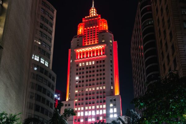 The Altino Arantes skyscraper in downtown Sao Paulo lit in red and orange at night between neighbouring buildings and palm fronds