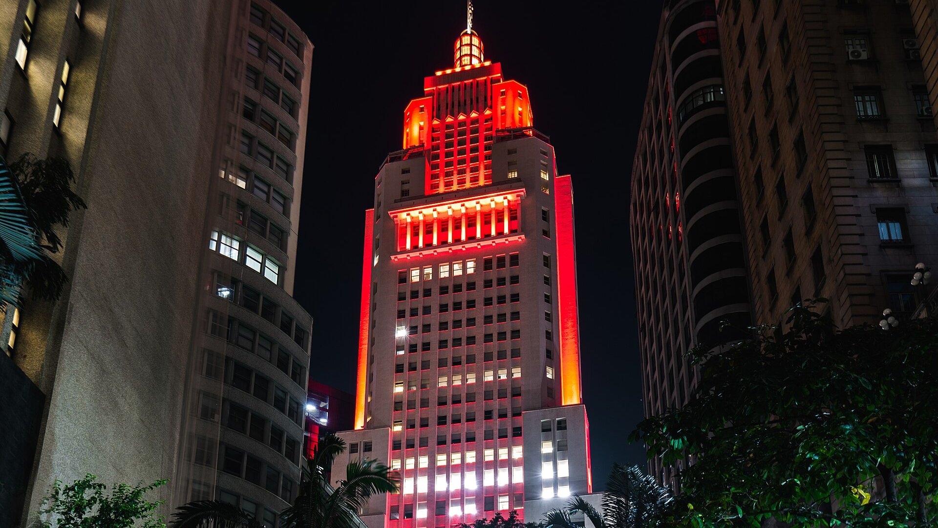 The Altino Arantes skyscraper in downtown Sao Paulo lit in red and orange at night between neighbouring buildings and palm fronds
