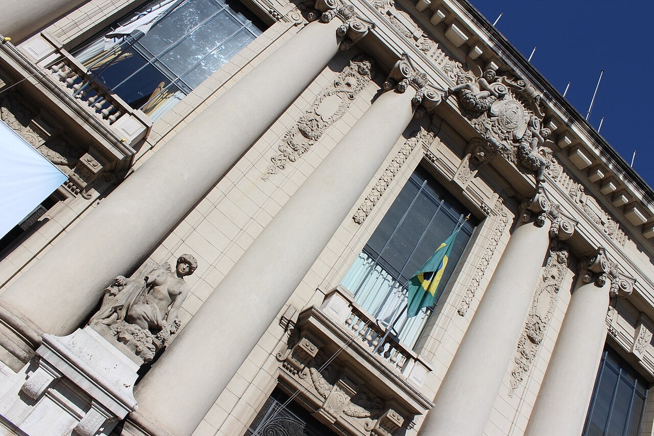 Tilted view of cream stone columns, balconies and a carved crest on the front of the Piratini Palace in Porto Alegre, with a Brazilian flag hanging from a window