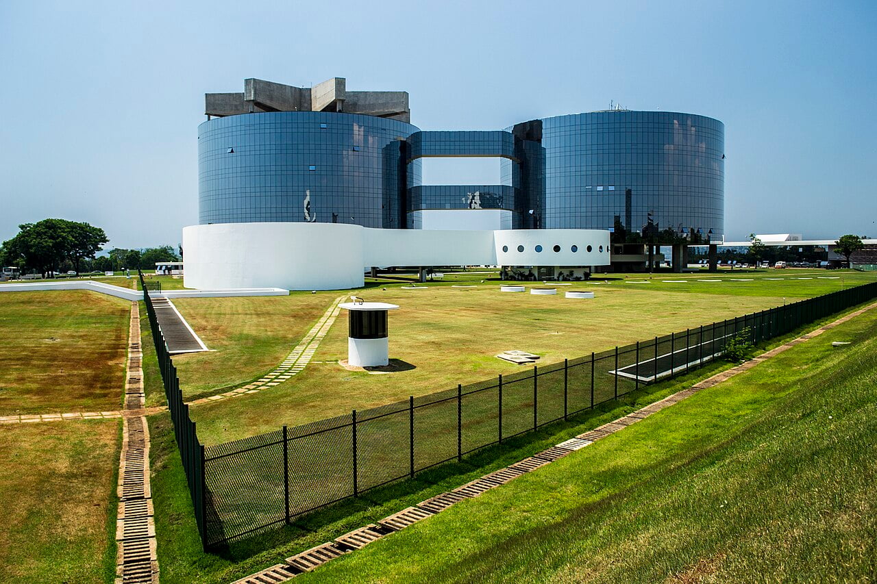 Two round mirrored-glass towers joined by glass bridges above a white curved base, on a fenced lawn in Brasília