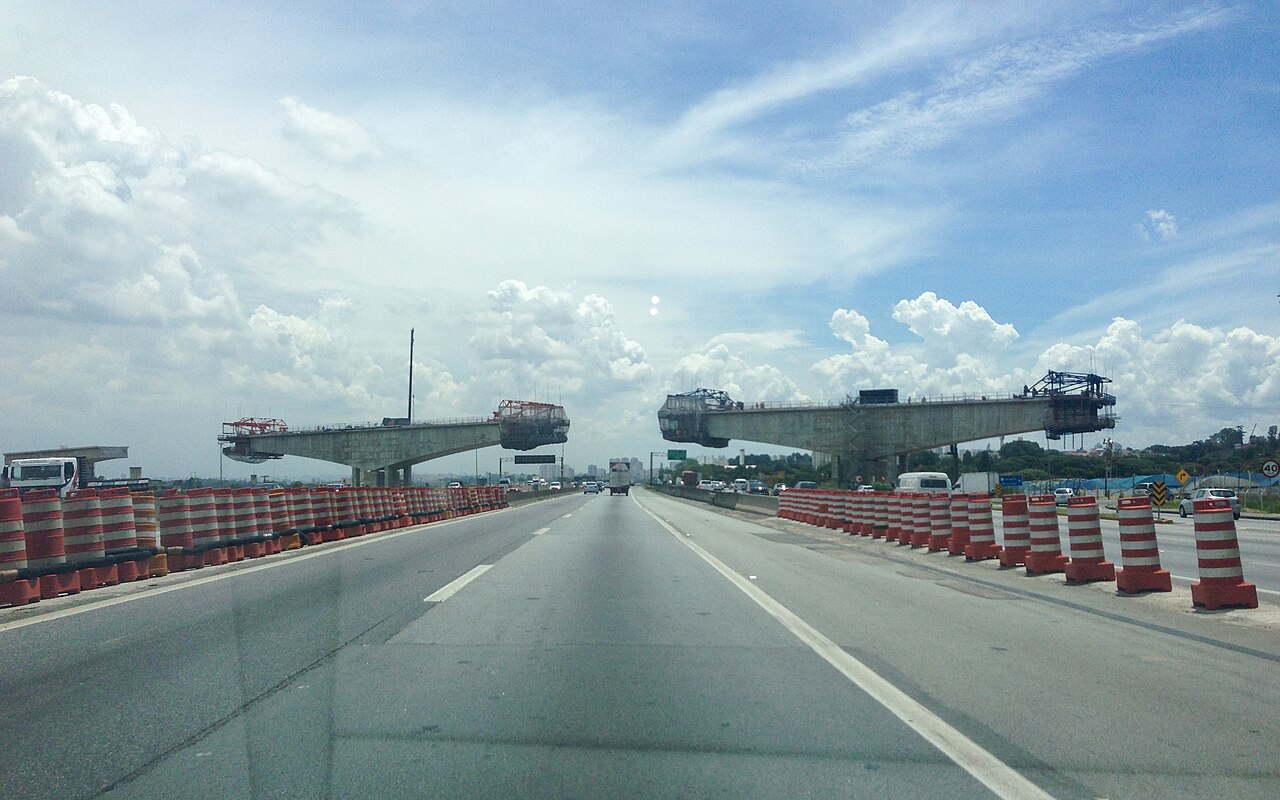 A multi-lane highway with orange and white traffic barrels on both sides and two unfinished concrete bridge spans over the road under a blue sky