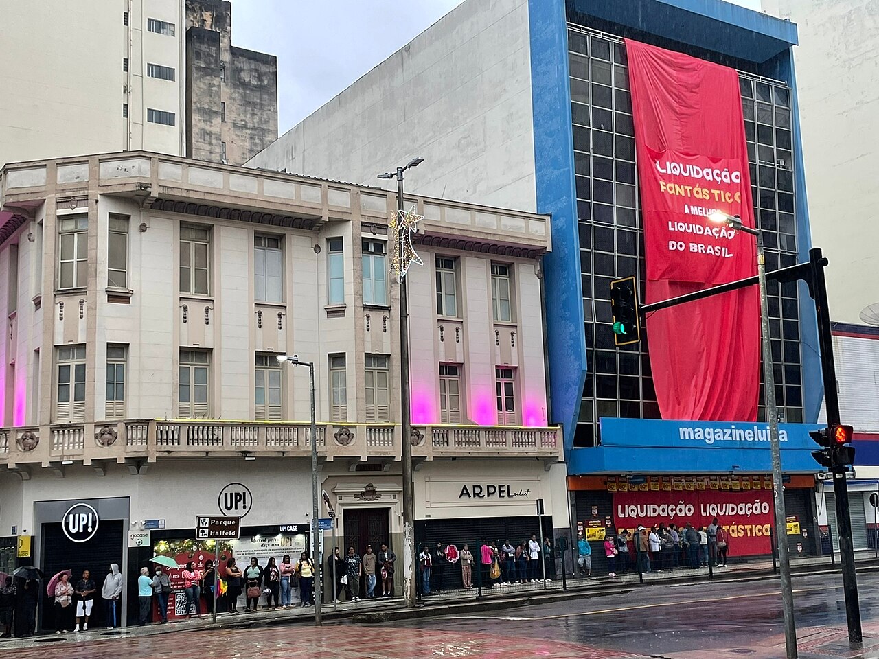 Shoppers queue on a rainy pavement outside a Magazine Luiza store whose blue facade carries a large red clearance banner