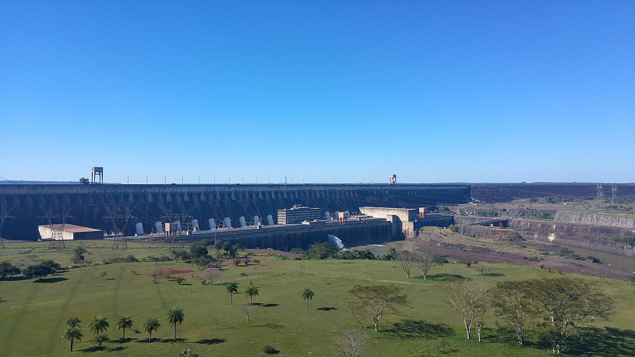 The long concrete wall of the Itaipu dam under a clear blue sky, with power pylons, a spillway jet and a grassy park in front