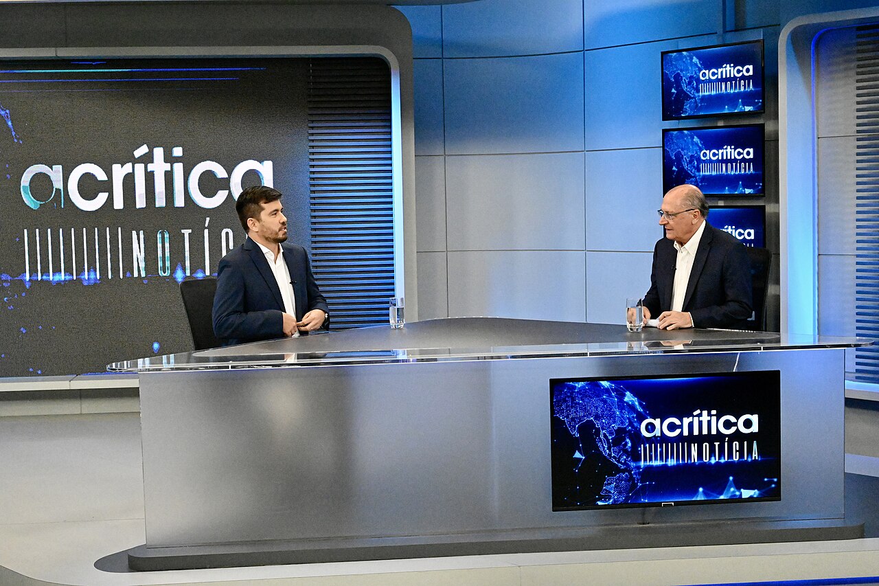 Geraldo Alckmin, in a dark suit and white shirt, sits at a TV studio desk facing an interviewer