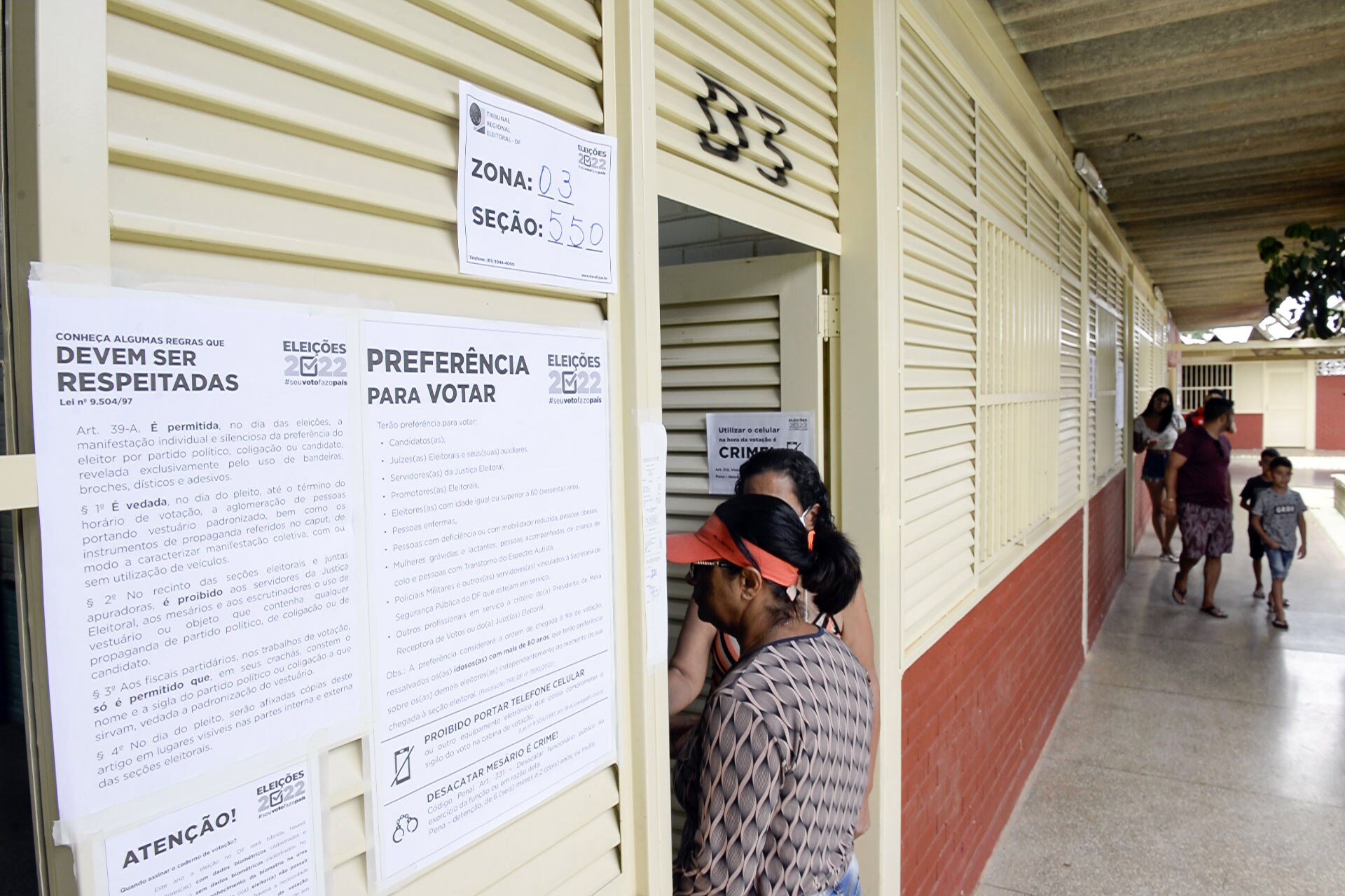 Signs on a polling-station door list the voting rules and priority-voting rights, with a voter waiting at the entrance, in Taguatinga Norte during Brazil's 2022 runoff
