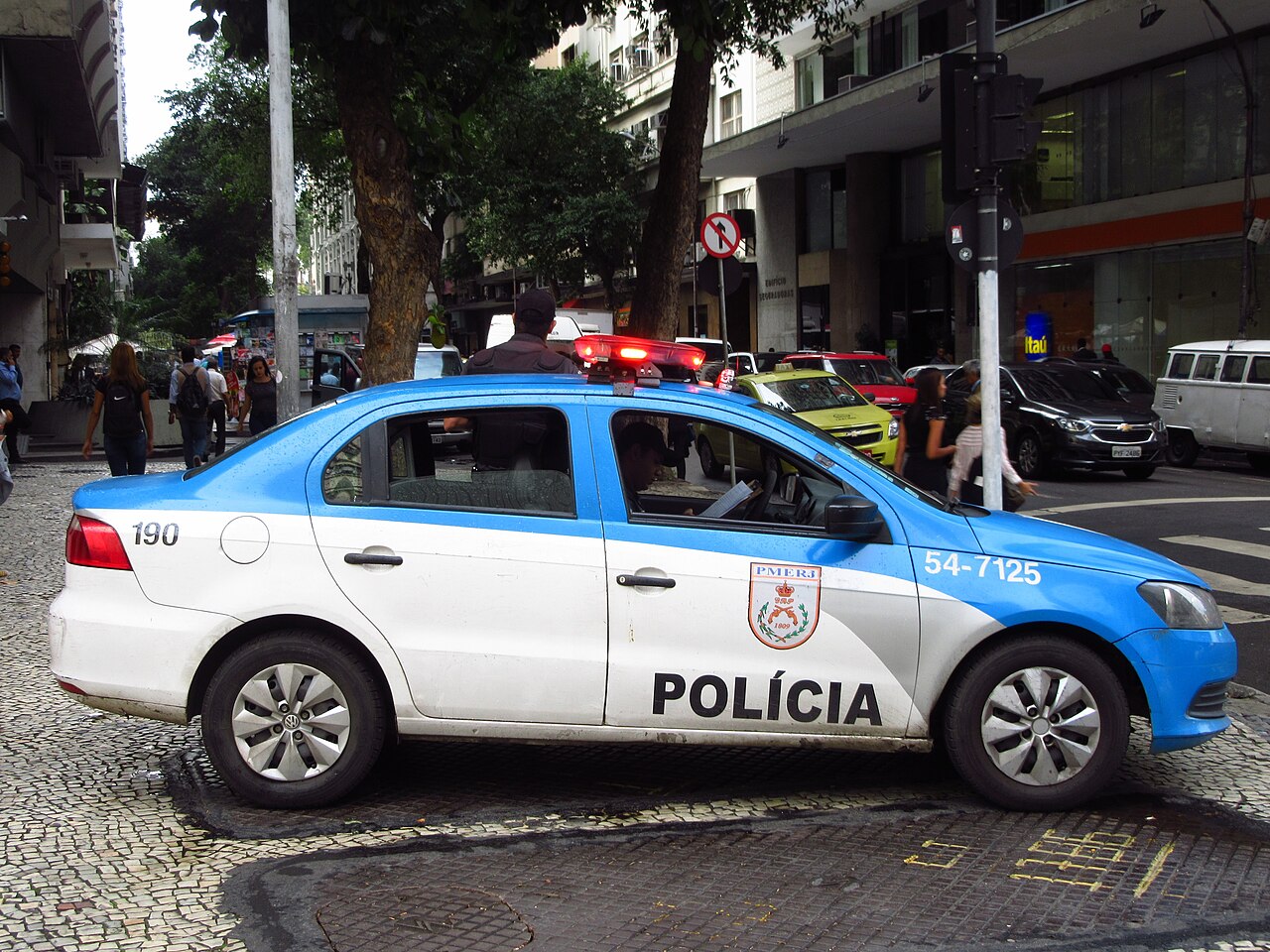 A white and blue Rio de Janeiro military police patrol car marked PMERJ and 190 parked on a mosaic pavement street.