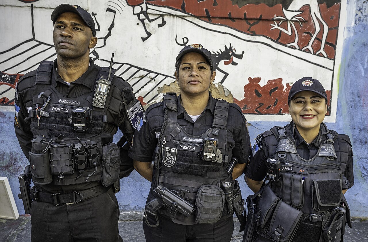 Three Rio de Janeiro military police officers in black tactical vests fitted with body cameras stand in front of a painted wall mural.