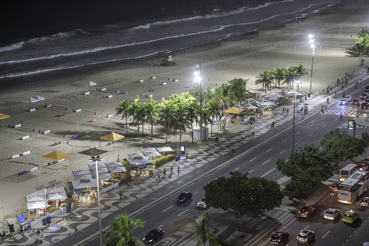 Copacabana beach and the wave-patterned promenade of Avenida Atlântica lit at night, with beach kiosks, palm trees and passing traffic.