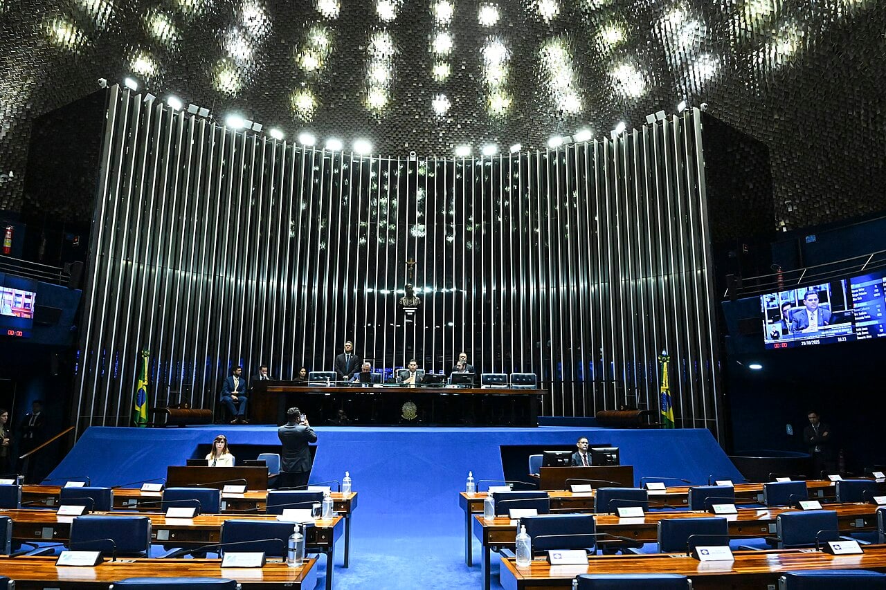 Brazil's Senate plenary chamber in Brasília with rows of wooden desks and blue chairs, mostly empty, below the presiding table and a wall of vertical metal tubes