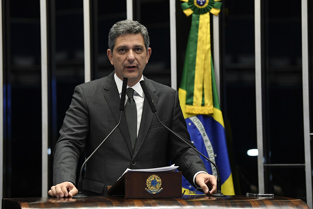 A grey-haired man in a dark suit speaks at a wooden lectern with two microphones, a Brazilian flag behind him