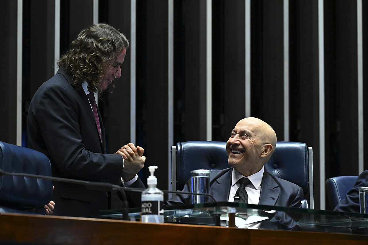 Two men in dark suits smile and talk at the presiding table of Brazil's Senate plenary, one standing with long curly hair and one seated