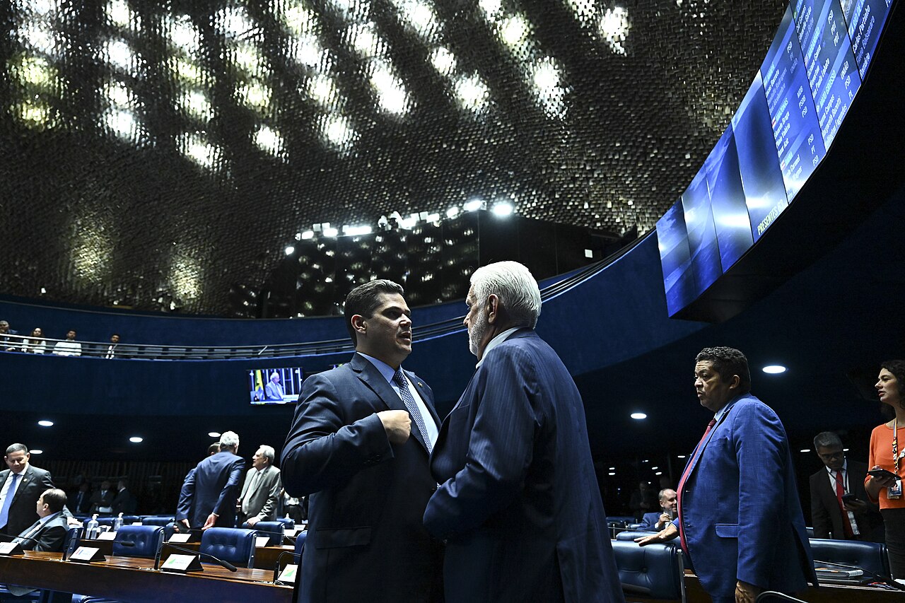 Two men in dark suits talk in the blue-lit plenary of Brazil's Federal Senate, with senators at their desks behind them