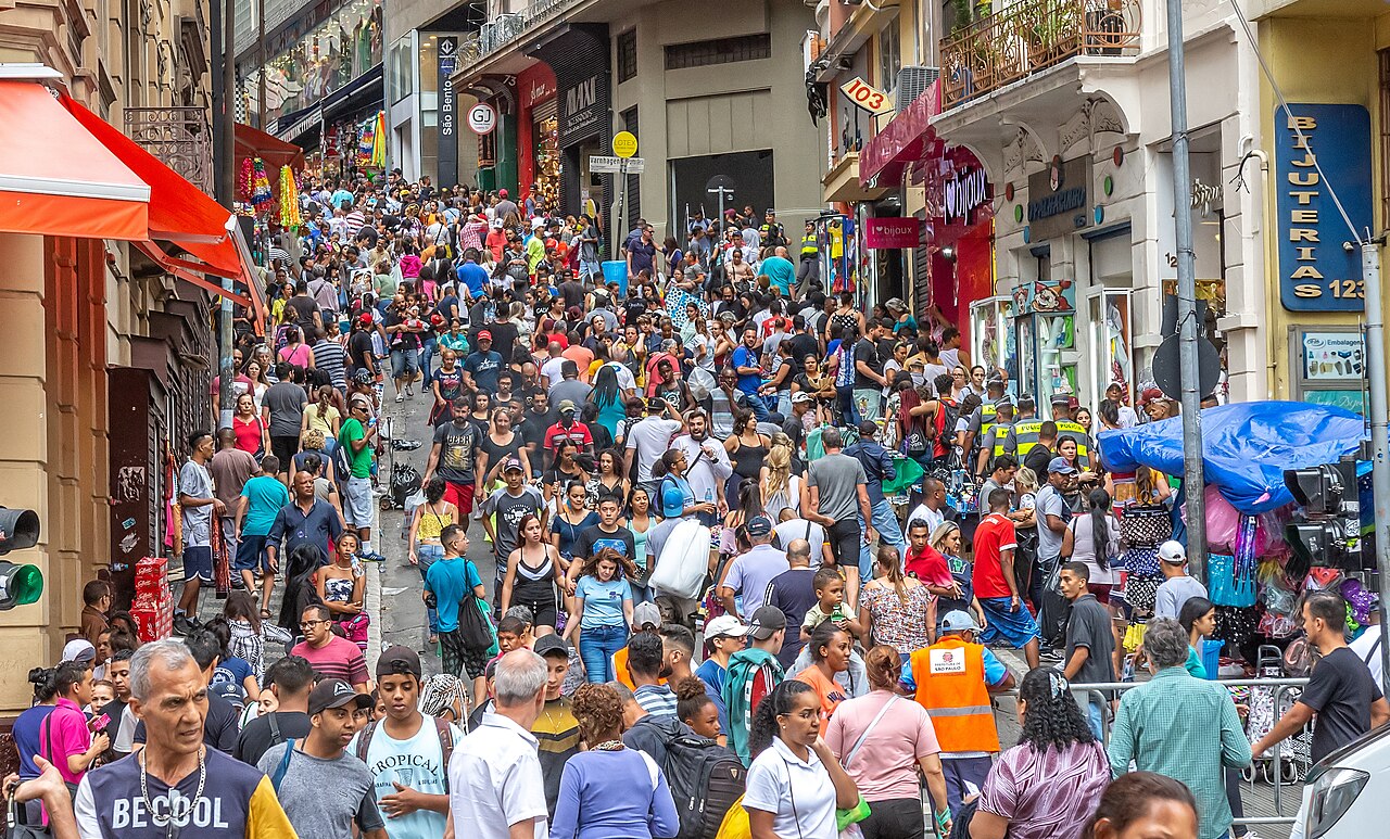 A dense crowd of shoppers walks up a steep, narrow street lined with small shops and market stalls in downtown São Paulo