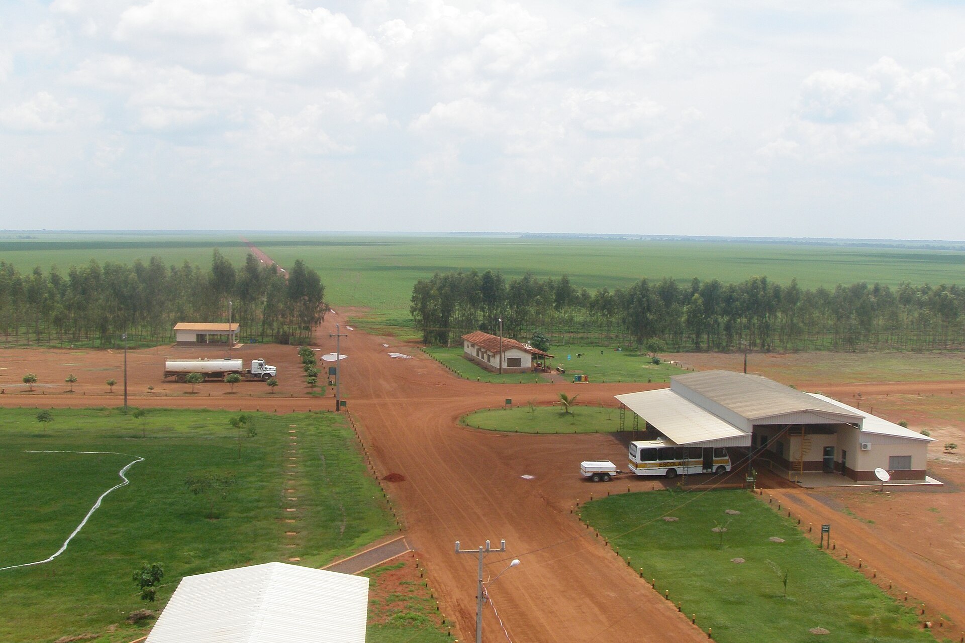 Aerial view of a soy farm in Mato Grosso, Brazil, with a red dirt road, farm buildings, rows of eucalyptus and green soy fields stretching to the horizon