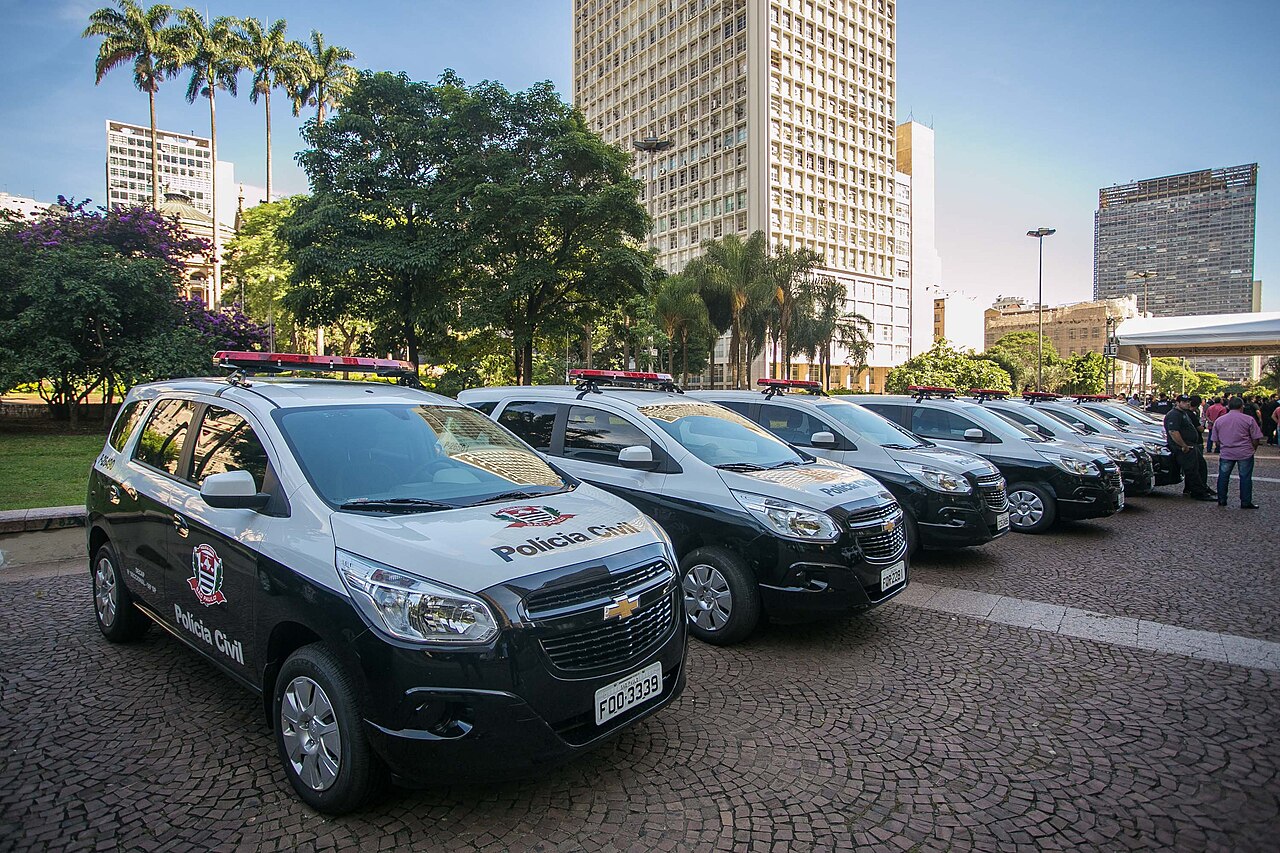 A row of black and white São Paulo Civil Police cars with red roof lights parked on a cobbled square in front of tall office towers
