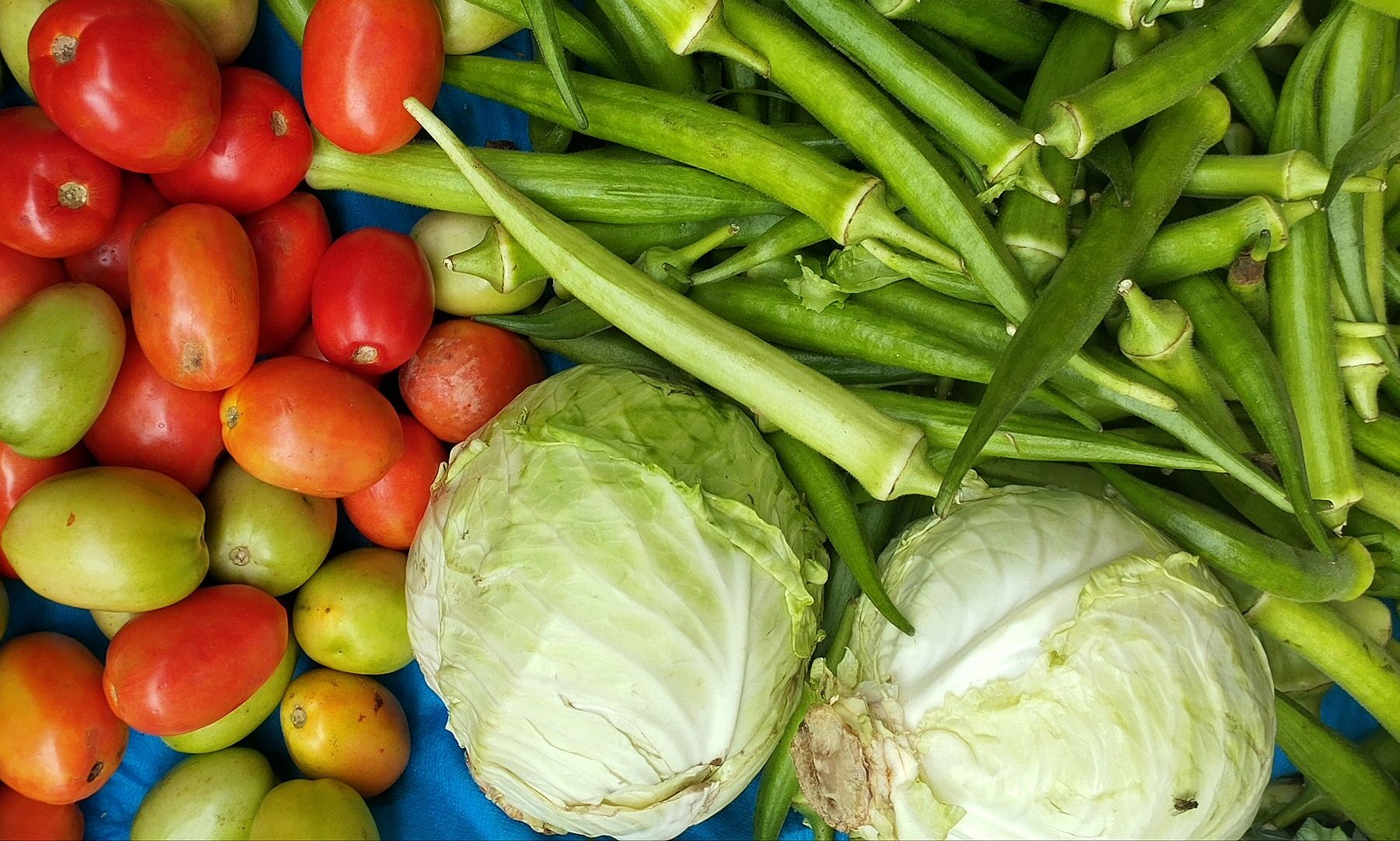 Red and green tomatoes, okra and two white cabbages laid out on a blue cloth at a street market stall