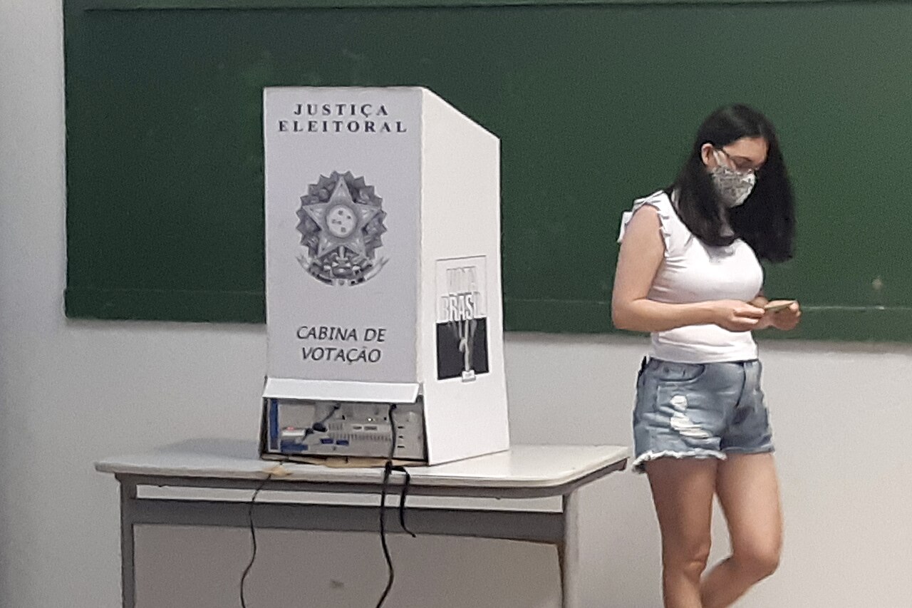 A young woman checks her phone beside an electronic voting booth marked Justiça Eleitoral in a Brazilian classroom