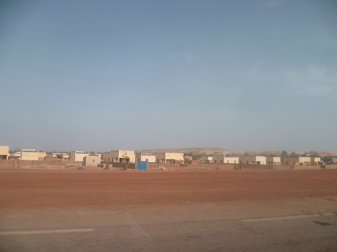 Rows of single-storey houses behind red earth, with a mine spoil hill on the horizon, at Sabsé in Burkina Faso