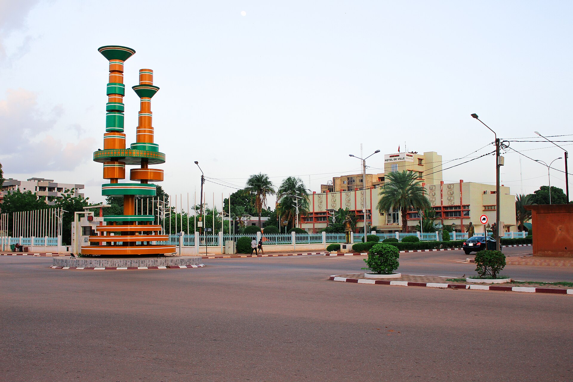 A roundabout monument and the Hôtel de Ville city hall in central Ouagadougou at dusk, with a few pedestrians and a car on the road
