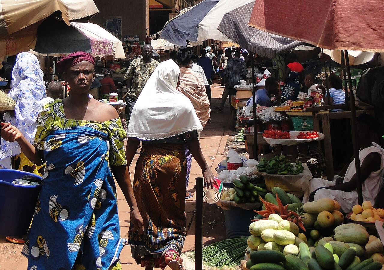 Shoppers walk between vegetable stalls and umbrellas in a covered market in Bobo-Dioulasso