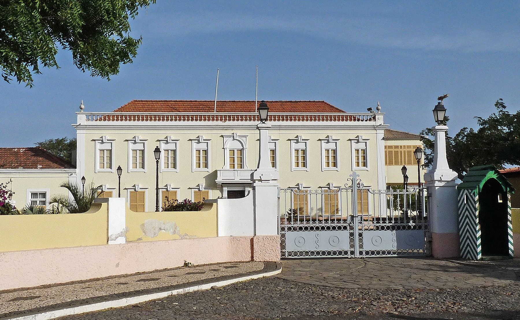 A cream-coloured two-storey palace with a red tiled roof behind a white iron gate, with a striped sentry box and a cobbled forecourt
