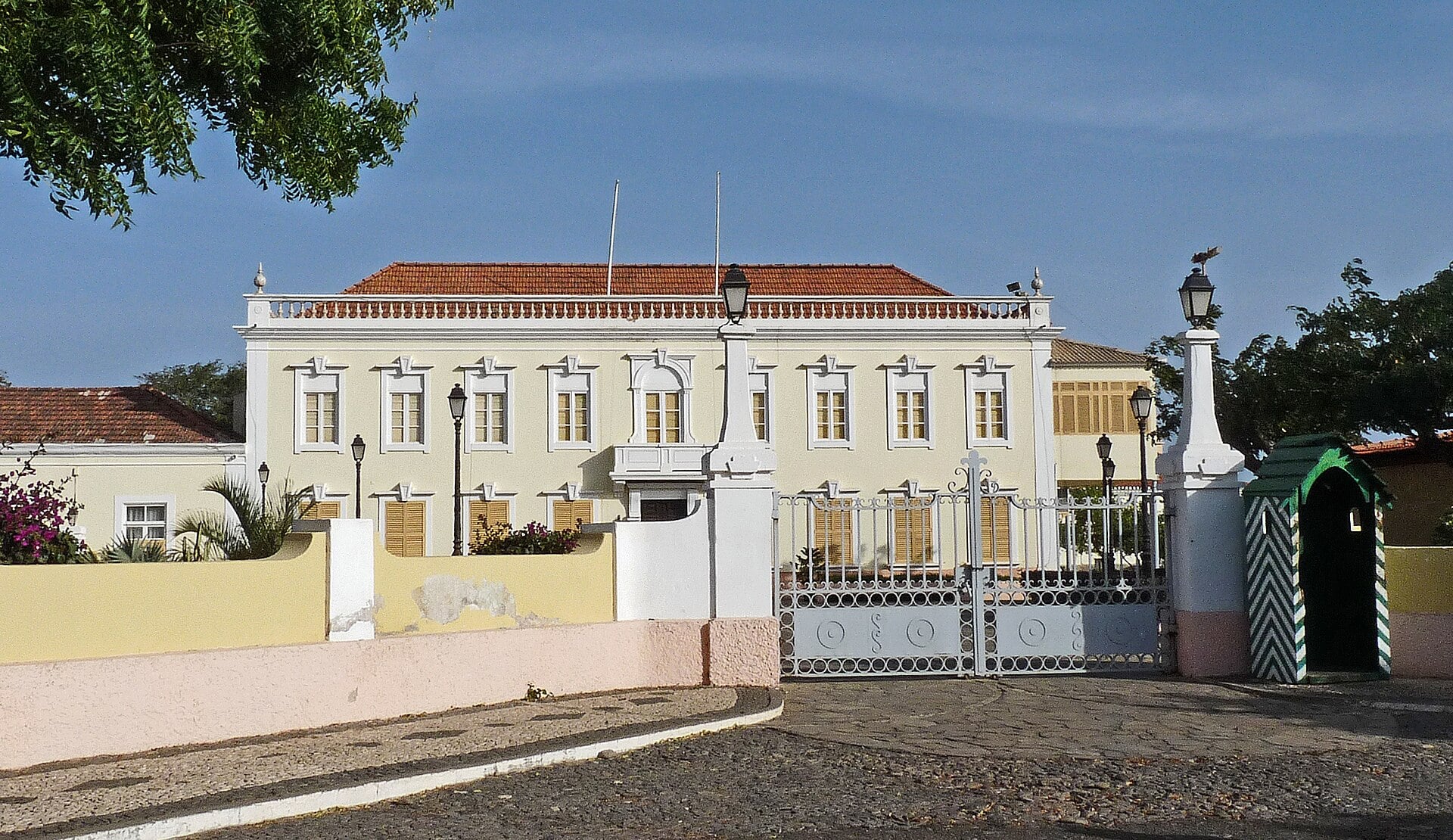A cream-coloured two-storey palace with a red tiled roof behind a white iron gate, with a striped sentry box and a cobbled forecourt