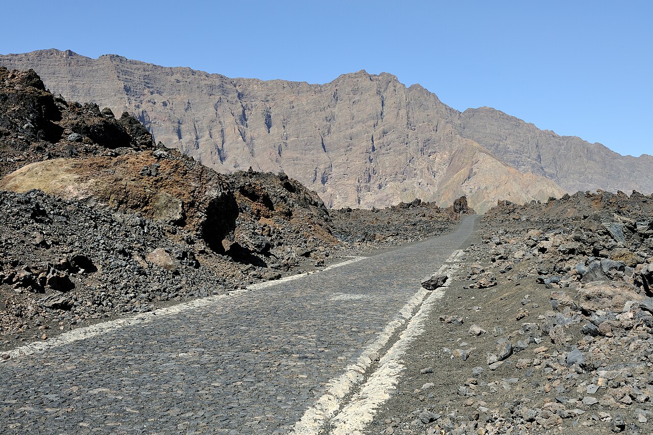 Cobbled road with white edge lines running through dark lava rock towards a high brown cliff wall under a blue sky on Fogo island
