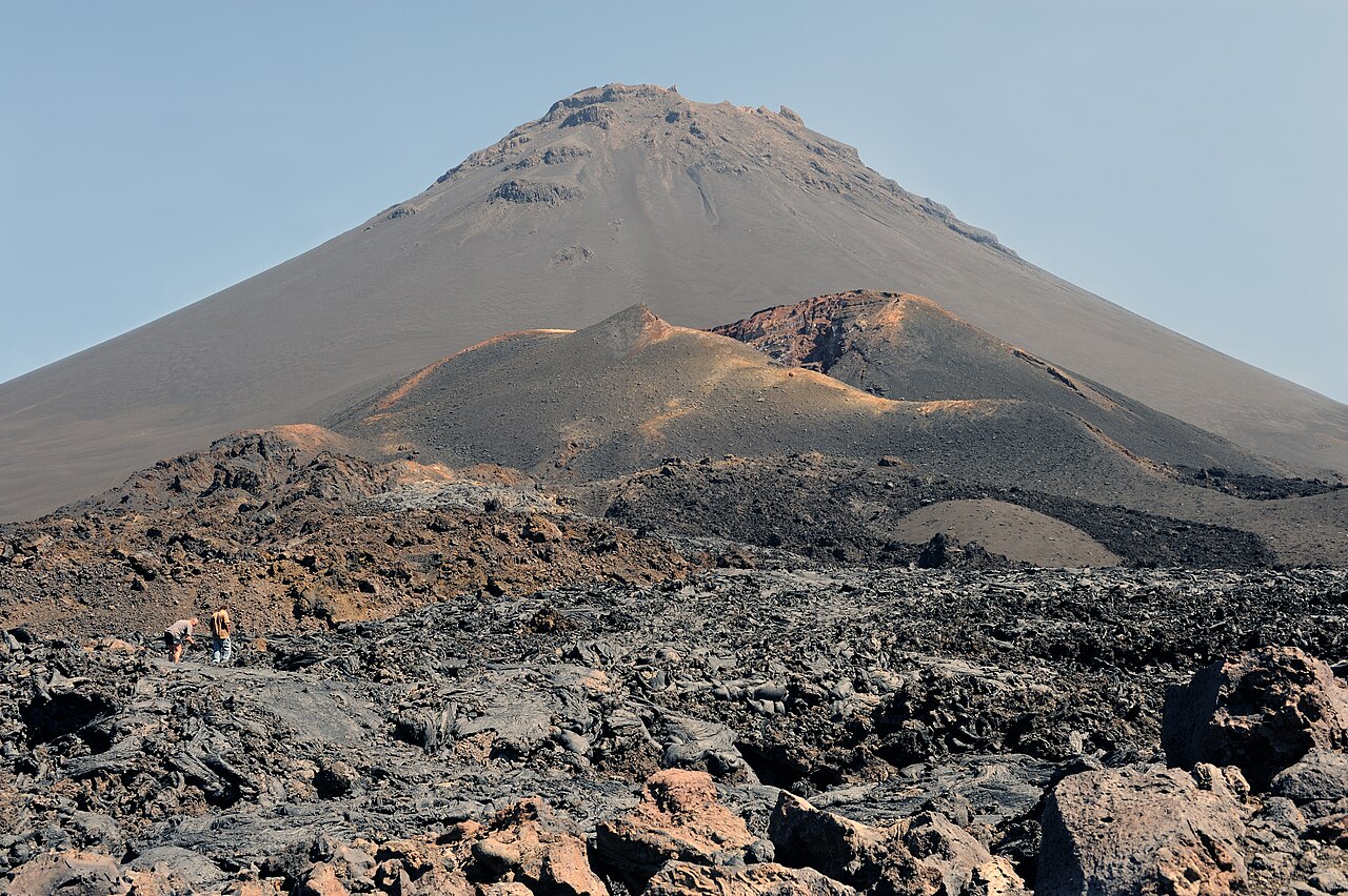 Cone of the Pico do Fogo volcano rising behind smaller craters and a dark lava field, with two people walking at left