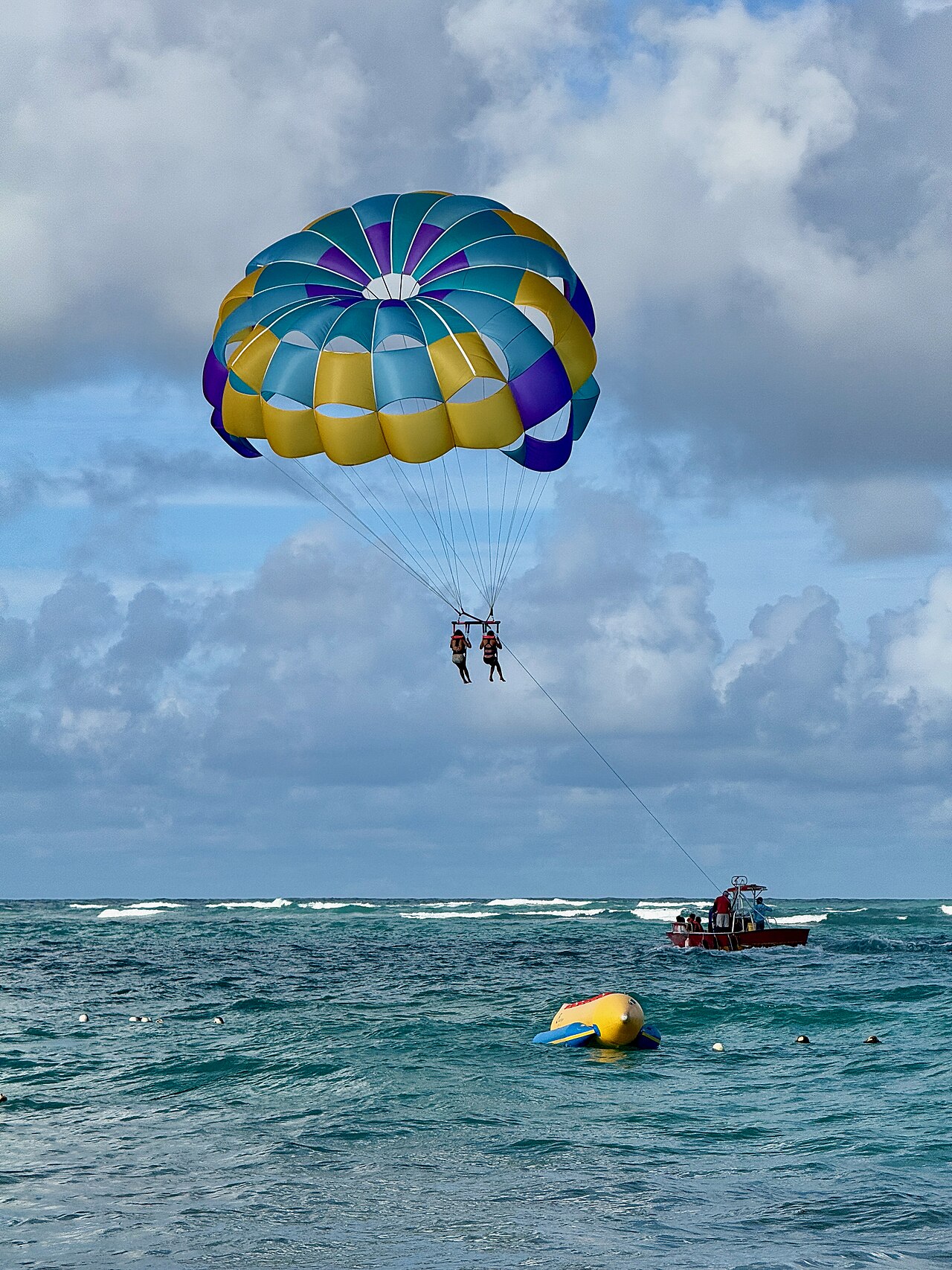 Two people parasailing under a blue and yellow canopy, towed by a red boat off Bávaro beach in Punta Cana.