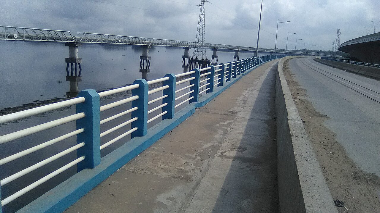 Two bridges crossing the wide Wouri river at Douala, with a blue-and-white railing in the foreground, a pylon and an overpass on the right.
