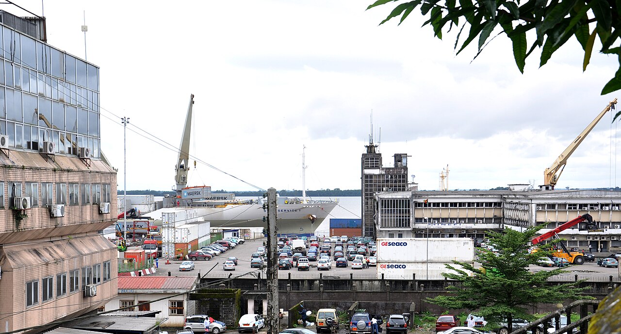 Container ship moored at a quay in the Port of Douala, with cranes, stacked containers, parked cars and port office buildings in the foreground.