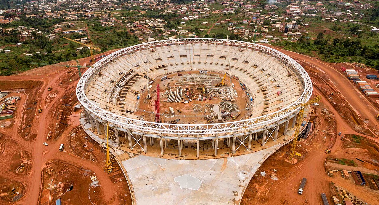 Aerial view of a large oval stadium under construction, with white roof trusses, cranes and red earth roads around it and houses beyond.
