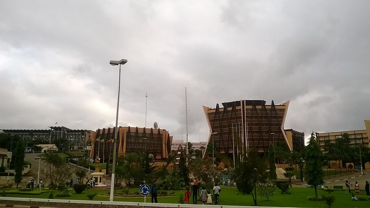 Two angular brown government office buildings stand among lawns and trees in Yaoundé under a cloudy sky.