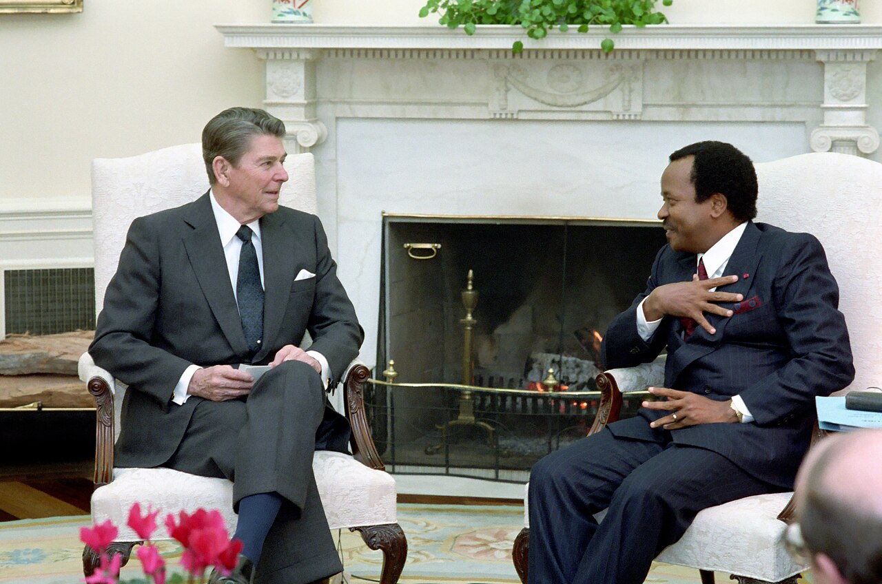 A younger Paul Biya sits in an armchair beside US President Ronald Reagan in front of the fireplace of the Oval Office.