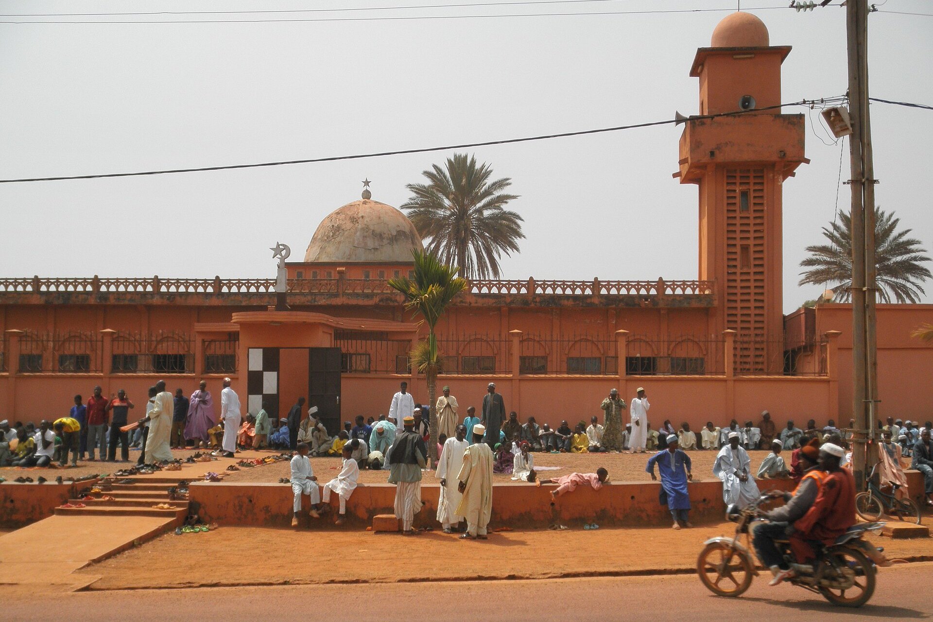 People in white and coloured robes sitting and standing outside the orange central mosque with its dome and minaret in Banyo, Cameroon