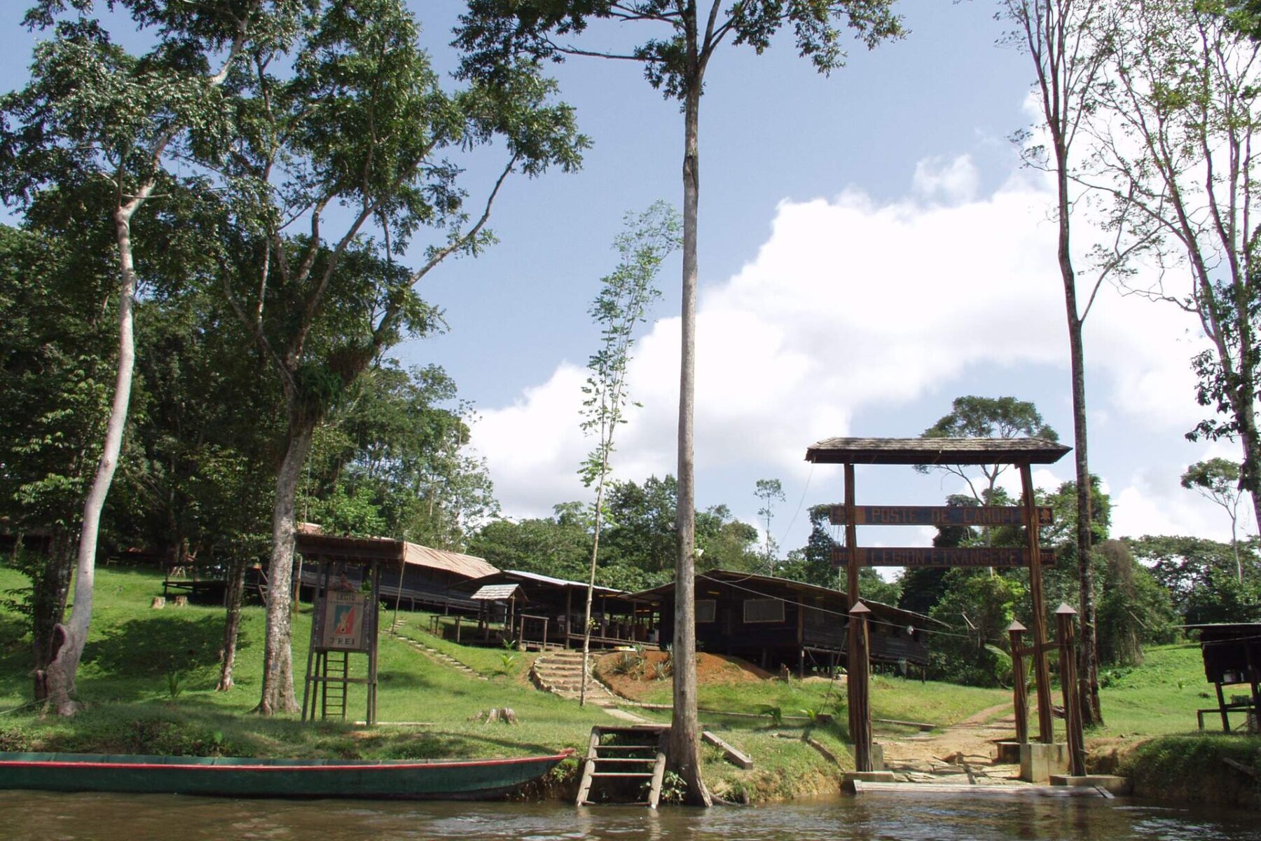 A wooden gateway and timber buildings on a grassy riverbank at Camopi, with a dugout canoe moored on the brown river