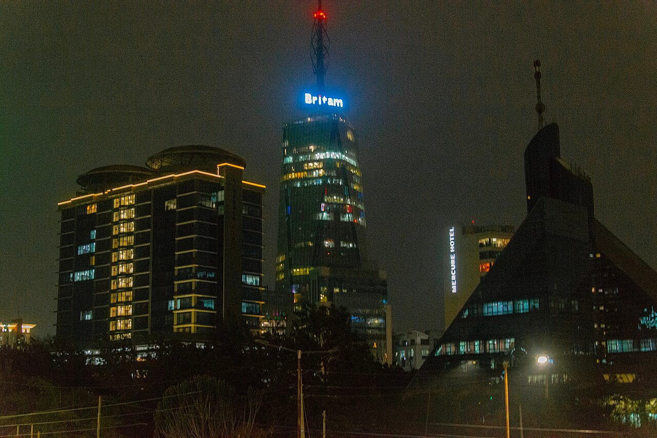 Britam Tower lit at night with its name in blue at the top, beside a lit apartment block, a Mercure hotel sign and a dark pyramid-shaped building in Nairobi