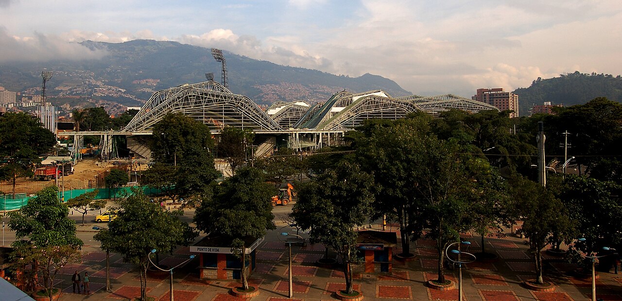Arched steel roofs of the indoor arenas at the Atanasio Girardot sports complex in Medellín, with a tree-lined plaza and green hills behind