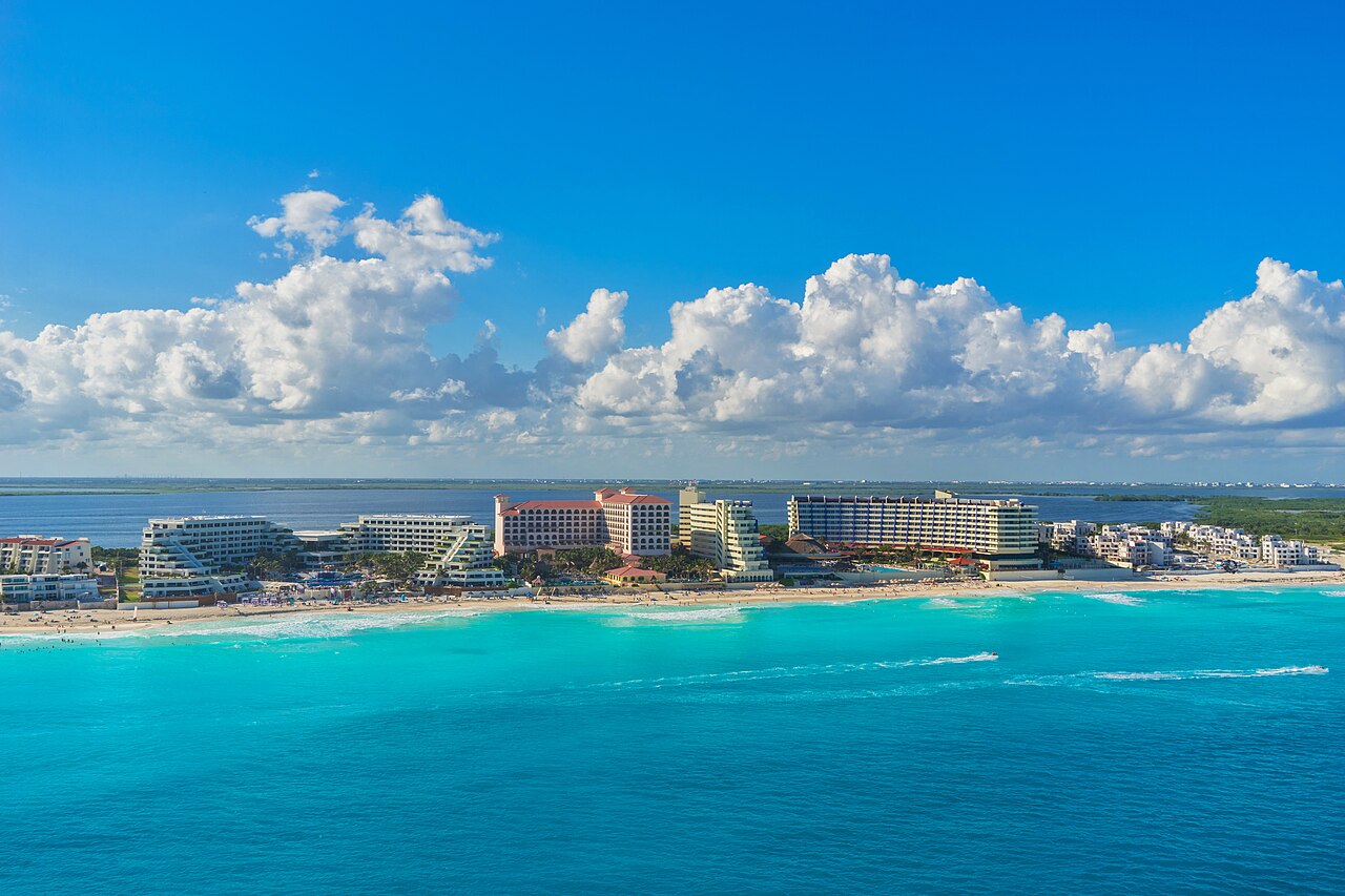 Resort hotels along the Cancún coast beside turquoise Caribbean water under a blue sky with large white clouds