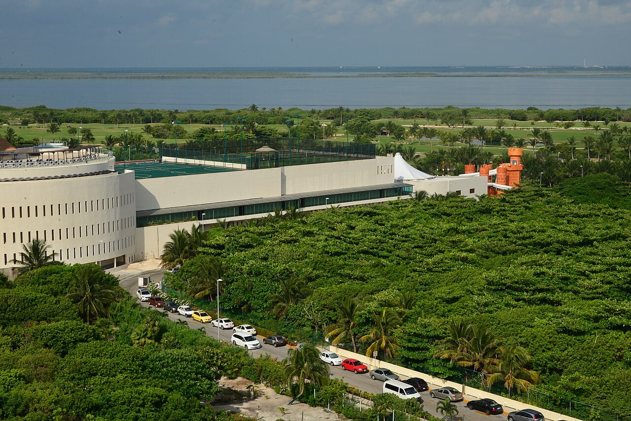 Resort buildings, tennis courts and green fairways in Cancún's Hotel Zone, with a wide stretch of calm water and a line of mangroves behind them.