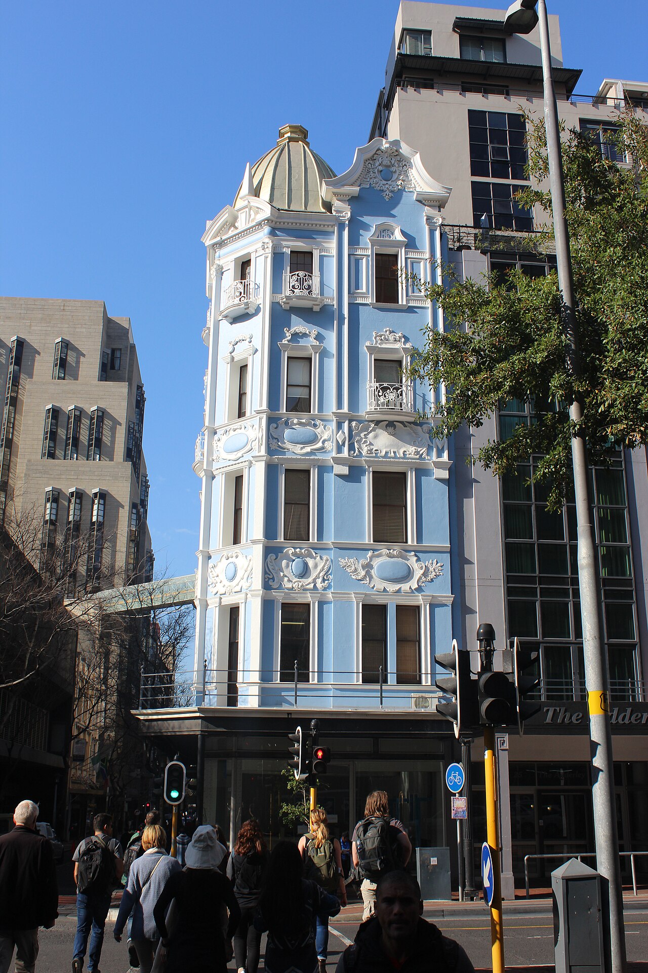 A narrow blue and white ornamental office building with a gold dome on a Cape Town street corner, with pedestrians at a crossing below