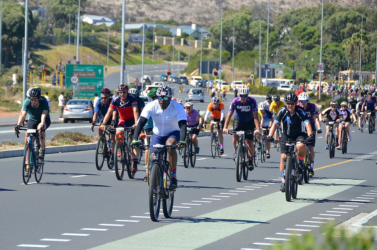 Riders in the 2024 Cape Town Cycle Tour near Sun Valley on the Cape Peninsula, with a road sign pointing to Simon's Town and Cape Point.
