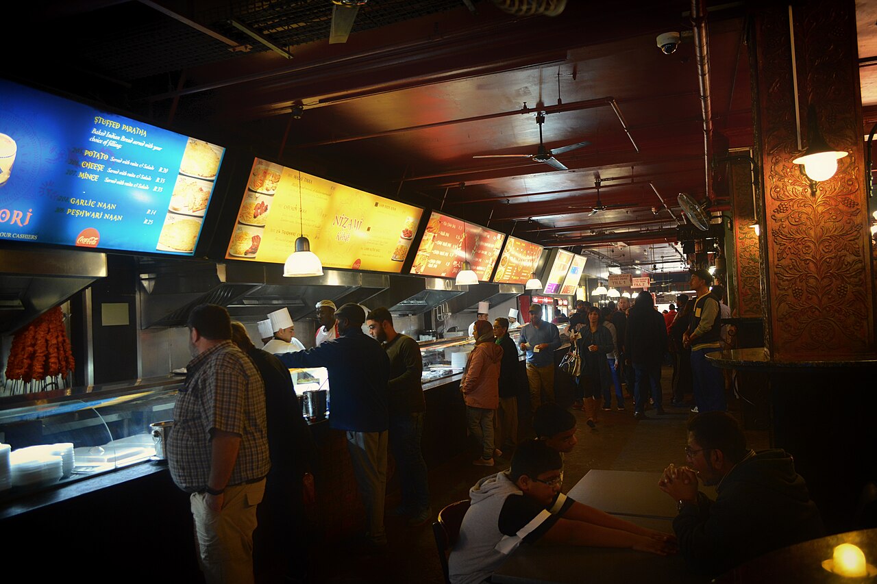 Diners queue at brightly lit food counters with menu boards at the Eastern Food Bazaar food hall in central Cape Town.