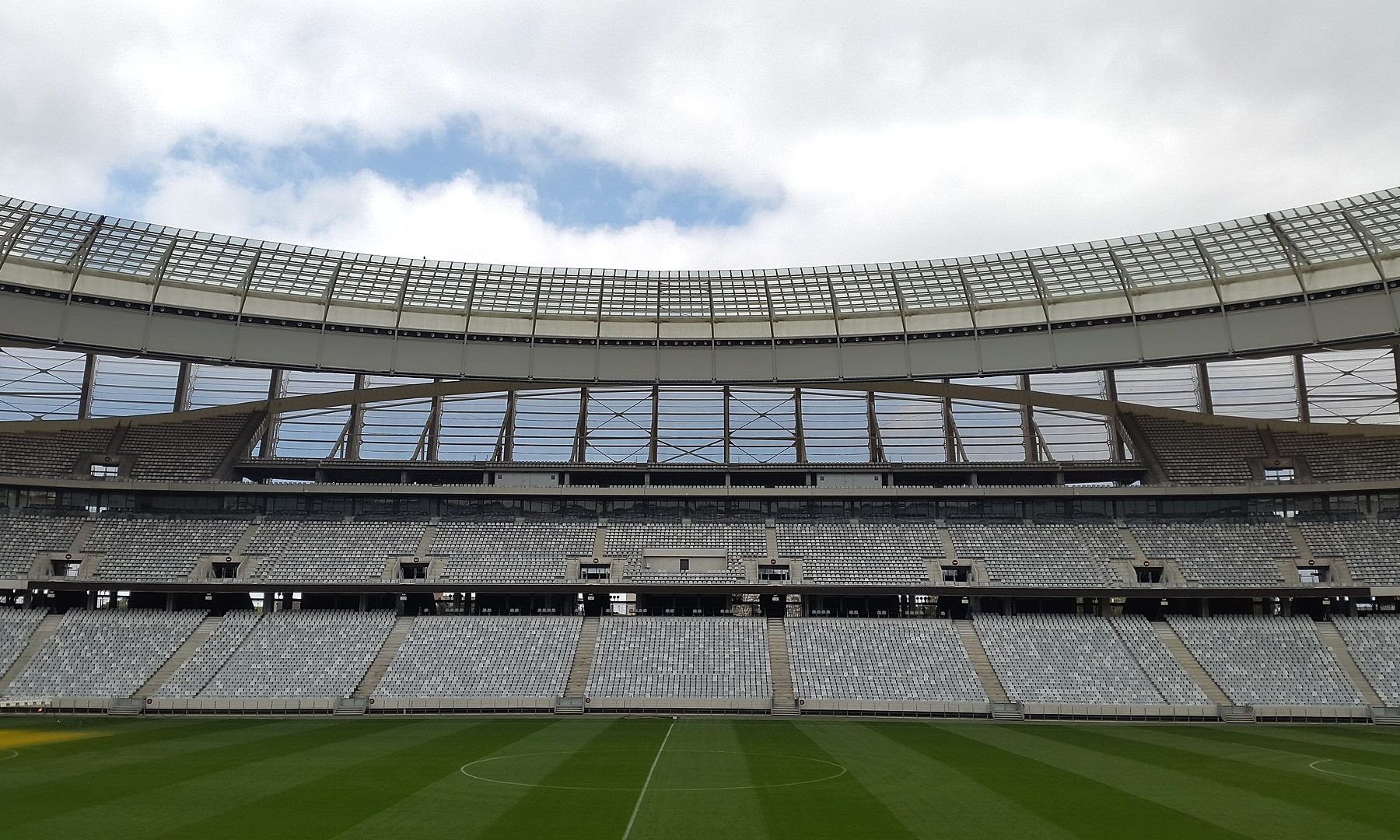 Empty lower and second-tier stands under the glass roof of DHL Stadium in Cape Town, with the pitch in the foreground