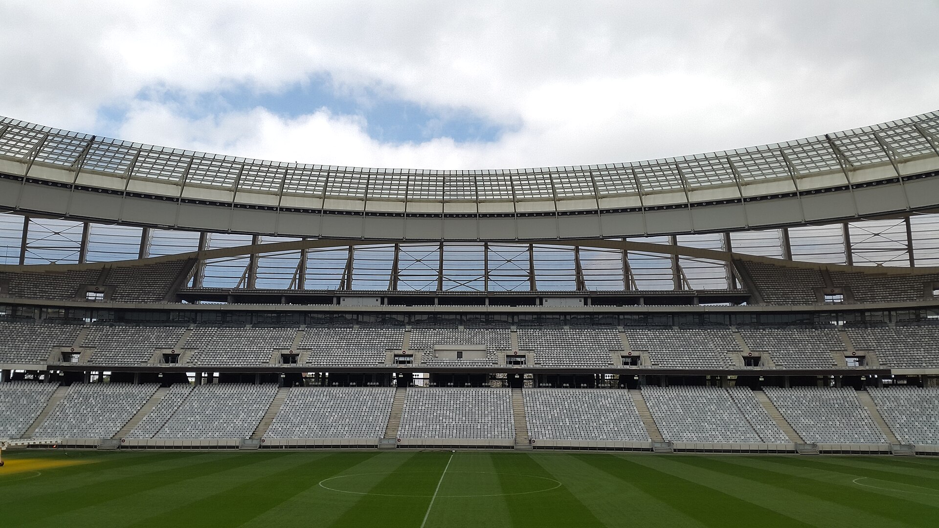 Empty lower and second-tier stands under the glass roof of DHL Stadium in Cape Town, with the pitch in the foreground