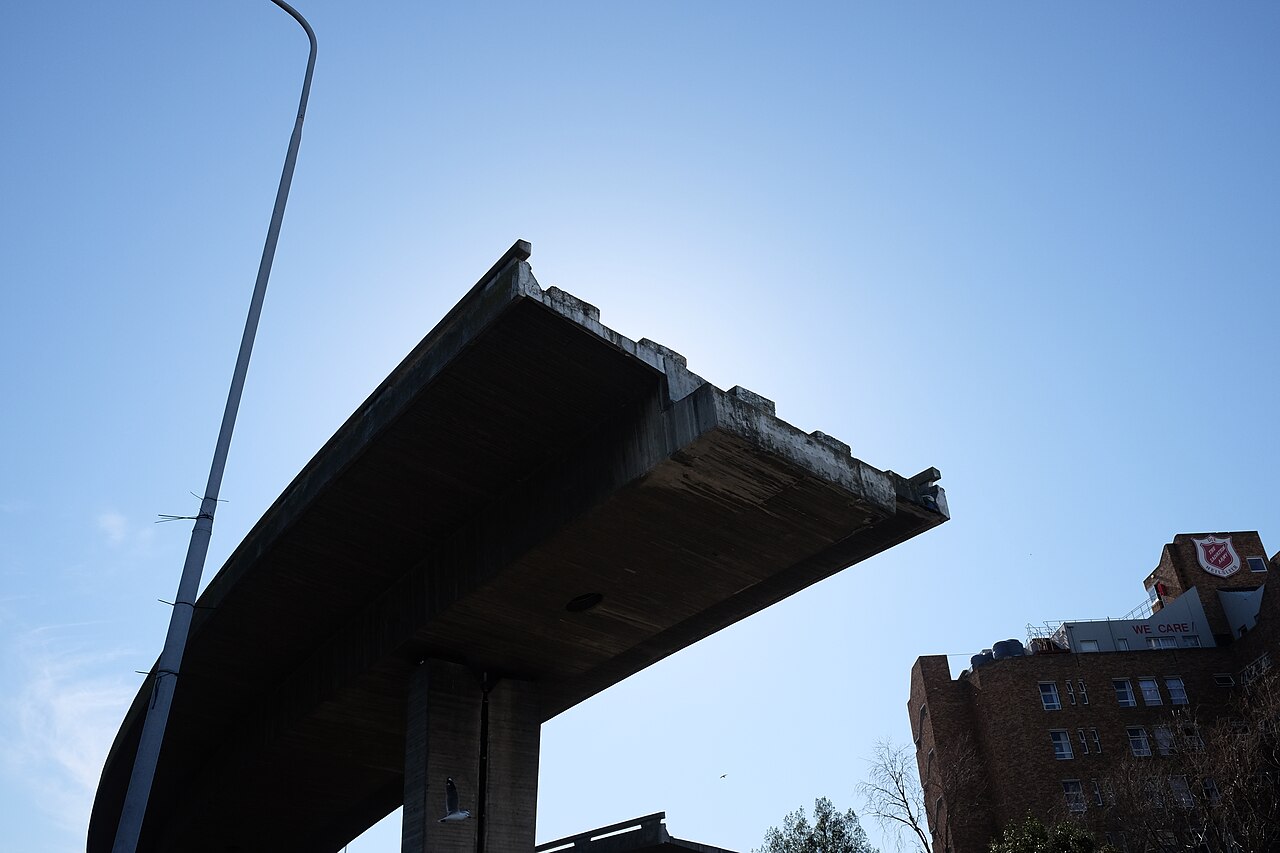 The underside of an unfinished concrete freeway deck in Cape Town, ending in mid-air against a blue sky