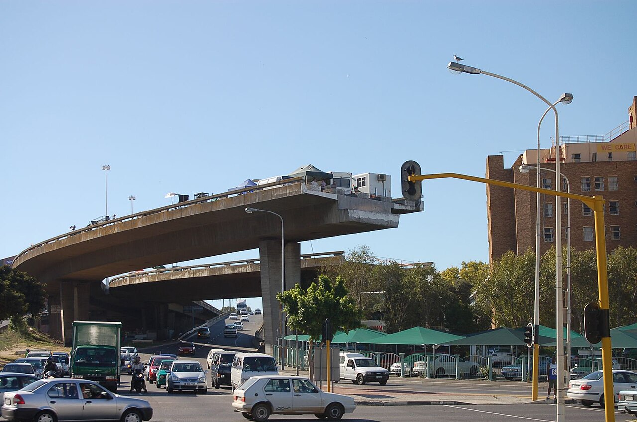 An unfinished elevated freeway in Cape Town ends abruptly in mid-air above a busy junction, with cars waiting at traffic lights below