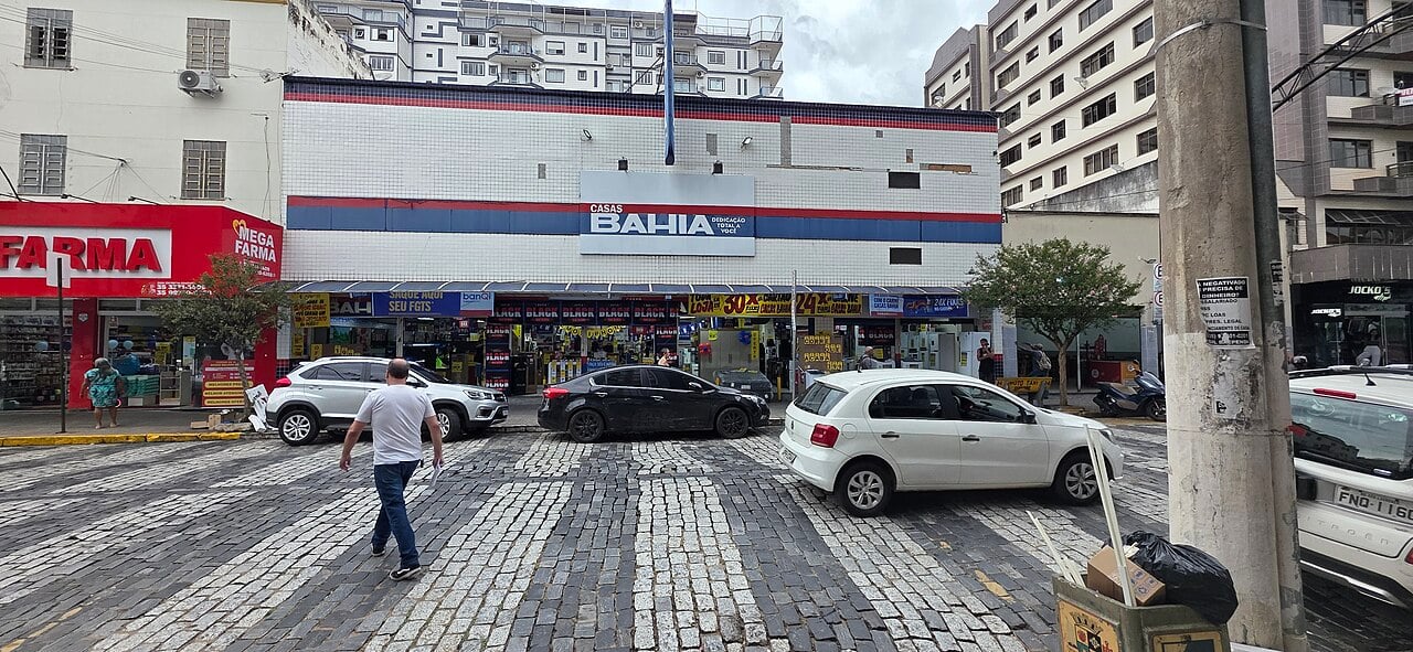 A Casas Bahia store with a white-tiled facade and blue and red stripes on a cobbled street corner, with parked cars and a pedestrian crossing in front