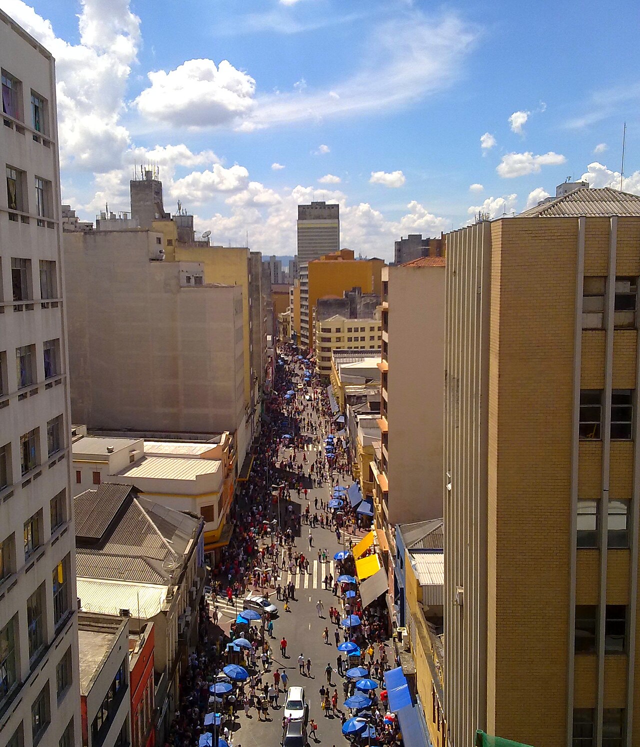 Aerial view down the crowded Rua 25 de Março in São Paulo, a shopping street packed with pedestrians and blue market umbrellas between tall buildings