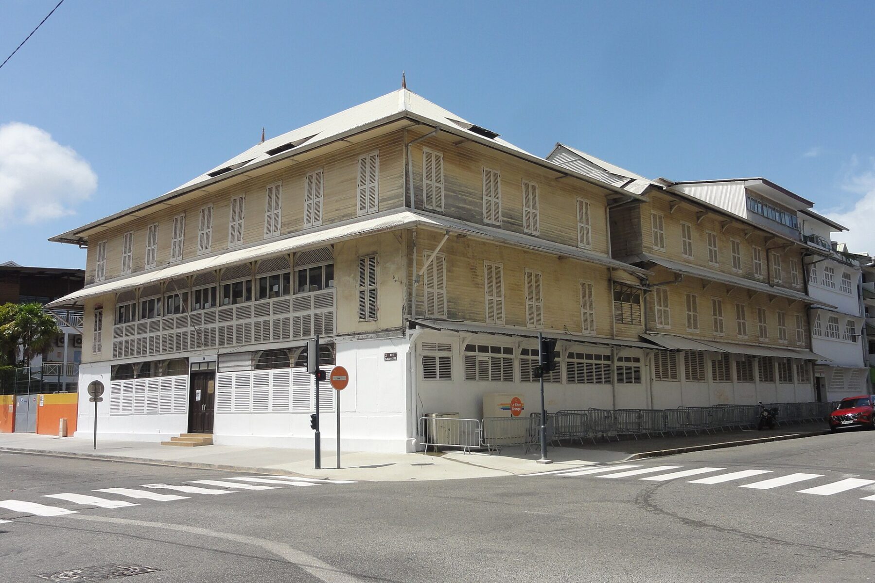 A three-storey wooden school building with louvred shutters and a white ground floor on a street corner in Cayenne, French Guiana, under a blue sky
